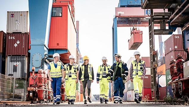 HHLA employees in protective workwear walk and talk across a Hamburg container terminal. In the background, a stacking crane transports a container.
