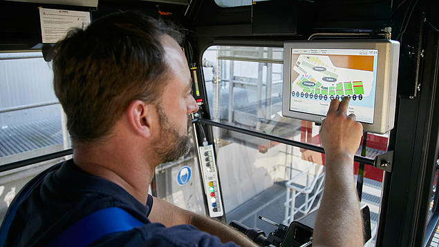 Ein Kranführer im Führerhaus bedient einen Touchscreen mit einer grafischen Benutzeroberfläche zur Terminalsteuerung; die HHLA entwickelt maßgeschneiderte Softwarelösungen für den operativen Alltag.