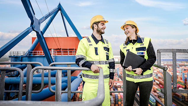 Two HHLA employees in safety gear stand on a container gantry crane, talking with a container terminal in the background.