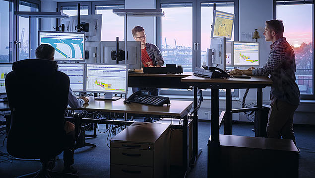 Employees of the Hamburg Vessel Coordination Center (HVCC) coordinate vessel arrivals at computer workstations with a view of the port.
