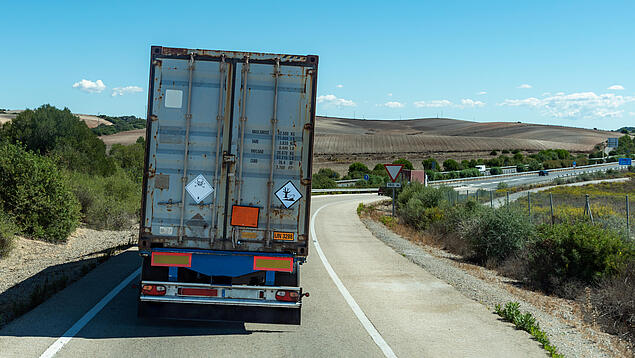 Rear view of a grey shipping container on a truck driving along a curving single-lane road. The container displays hazardous material placards, including a skull and crossbones (toxic) and a dead fish/tree symbol (environmental hazard). The background features rolling brown hills, a clear blue sky, and a highway junction with road signs in the distance.
