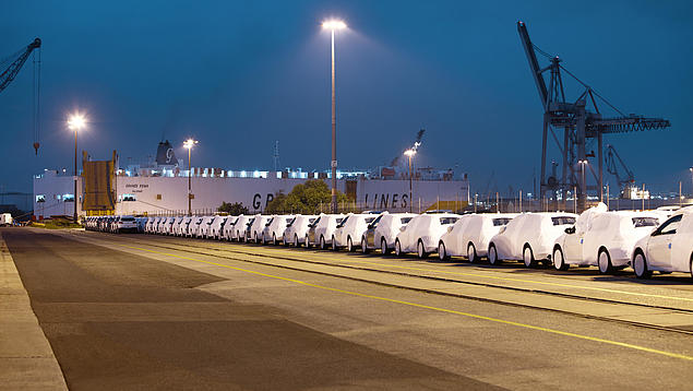 Lagerung von RoRo-Ladung am Unikai Hamburg bei Nacht: Lange Reihen weiß folierter Neufahrzeuge stehen auf einer beleuchteten Außenfläche vor einem großen Autotransportschiff (RoRo-Carrier). Im Hintergrund sind Hafenkrane und die Schiffsrampe erkennbar.
