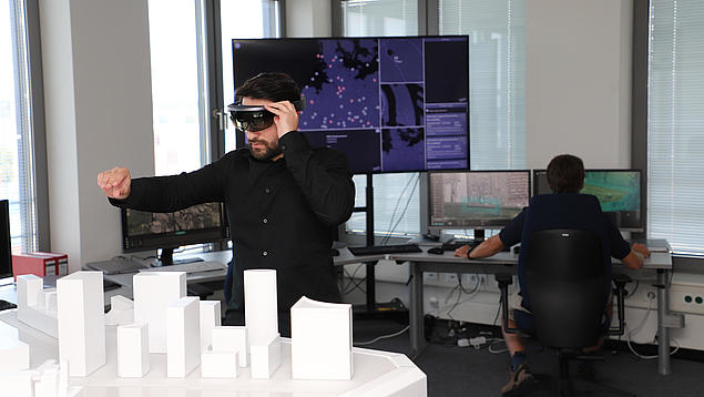 Ein HHLA-Spezialist trägt eine Mixed-Reality-Brille und interagiert über einem physischen Architekturmodell mit digitalen 3D-Daten; im Hintergrund sind weitere Planungsmonitore zu sehen.
