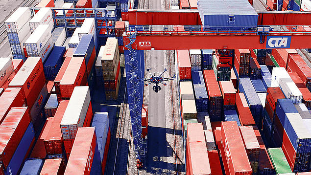An industrial drone by HHLA Sky flies over the block storage yard at the HHLA Container Terminal Altenwerder (CTA) in Hamburg, passing between stacking cranes and stacked containers.
