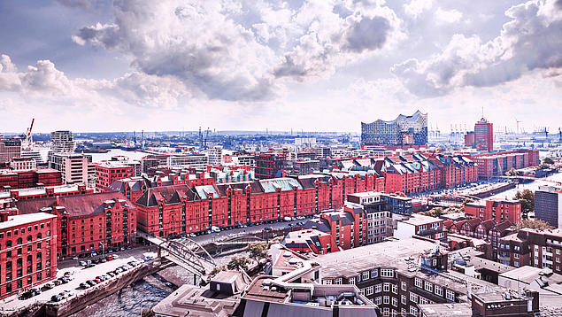 Panoramic view of Hamburg's Speicherstadt with red brick warehouses along a canal, the Elbphilharmonie and harbour cranes visible in the background.